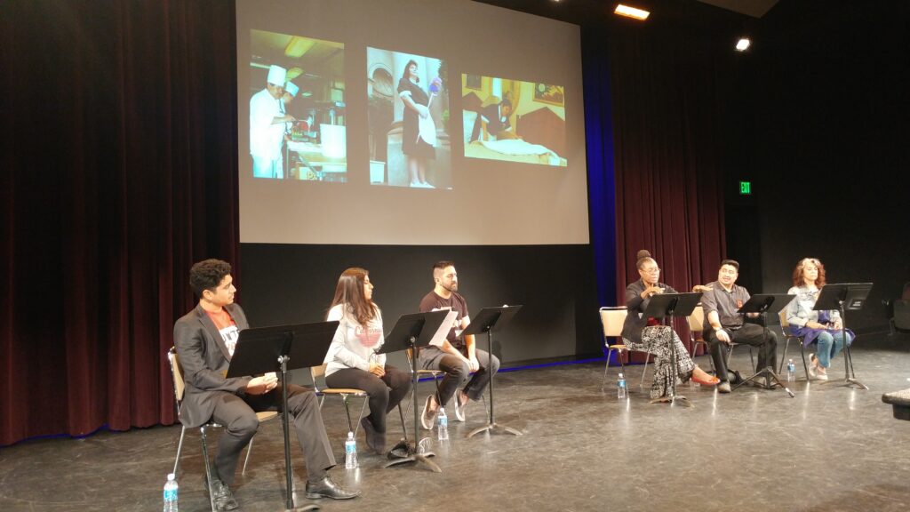 Six actors, 3 male presenting, 3 female presenting, sit on a stage with music stands in front of them. Behind them is a large projection screen with images of hospitality workers on it.