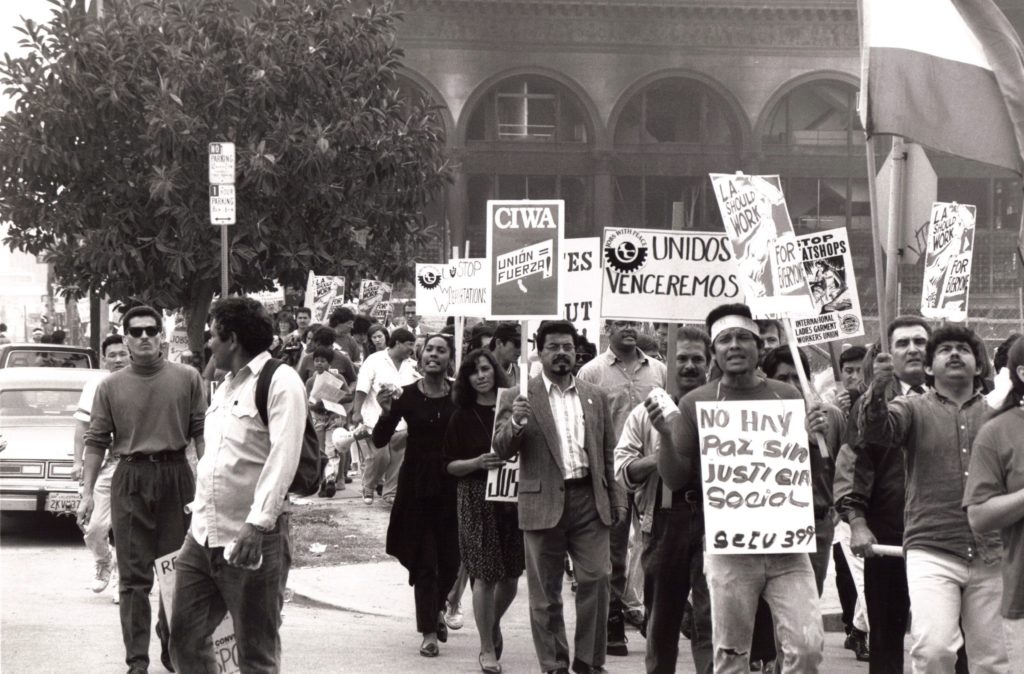 Black and white image of a demonstration with many people cheering. They hold signs representing many organizations, including: "Unión = Fuerza" (from CIWA), "Unidos Venceremos" (Jobs With Peace", "Stop Sweatshops" (ILGWU) and "L.A. Should Work for Everyone" (Justice for Janitors).