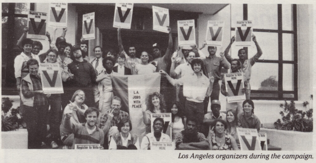 Grainy black and white photo of a group of young (ish) men and women. IN the center, they hold a banner reading "L.A. Jobs with Peace", Many others in the photo are displaying signs with a large "V" on them (signs also say "Register to Vote here") or making a V sign with their fingers. They are posing outside the entrance of a building. IN the bottom corner, the photo is captioned "Los Angeles organizers during the campaign."