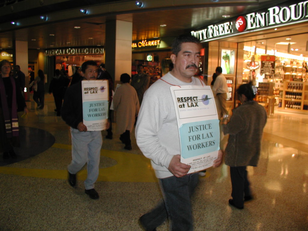 Slightly blurry color image of workers picketing inside an airport terminal. The men wear signs reading "Respect at LAX - Justice for LAX Workers." The Duty Free store can be seen in the background.