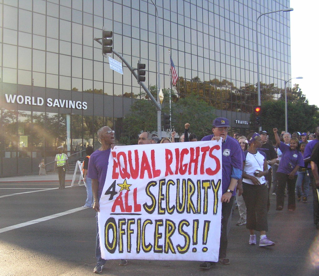 Workers in purple t-shirts carry a banner reading "Equal rights 4 All Security Officers!!" lead a crowd of demonstrators marching in the street. Behind them is a glass office tour with a "World Savings" logo. A police officer directs traffic. Sunbeams break around the edge of the left hand side of the frame.