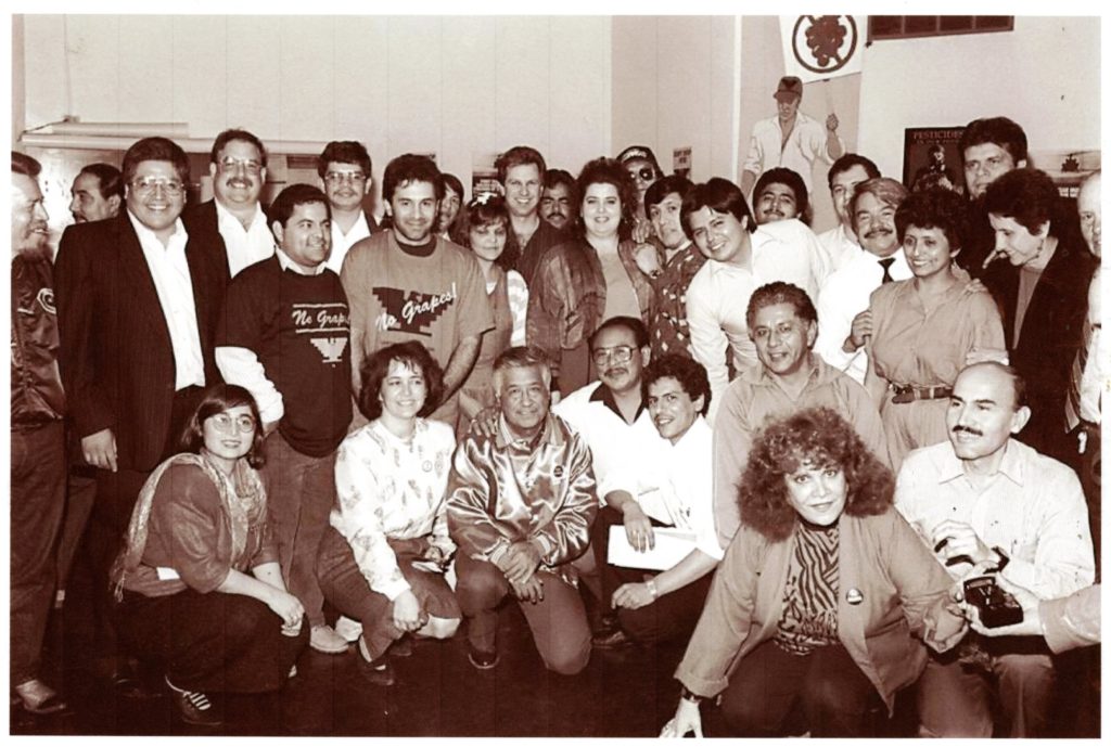 Black and white image shows a group of Latinx workers gathered around Cesar Chavez, who is kneeling wearing a silken jacket. To his right is Maria Elena Durazo. Everyone is smiling enthusiastically.