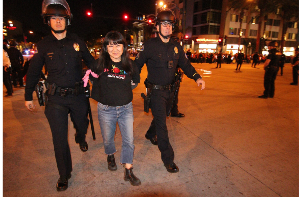 Two white police officers in riot gear escort a woman away from an action where many more police are lined up in the street. She is smiling, wearing jeans and boots and a UNITE-HERE t-shirt.