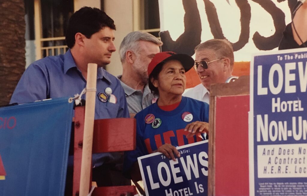 Labor leaders chatting on stage during a rally. In the middle is Dolores Huerta, wearing a United Farmworkers hat, holding a picket sign that says "LOEWS Hotel is Non-Union"