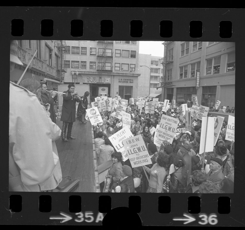 Black and white image appears as a negative, with perforated strips on top and bottom. It depicts a group of workers in an alley outside a garment factory with a visible marquee reading "Dresses." The workers carry signs reading "IBEW joins ILGWU to Keep Americans Working" and "Stop Exporting Jobs". Mariachi musicians play trumpets and violins for the crowd from a stage.