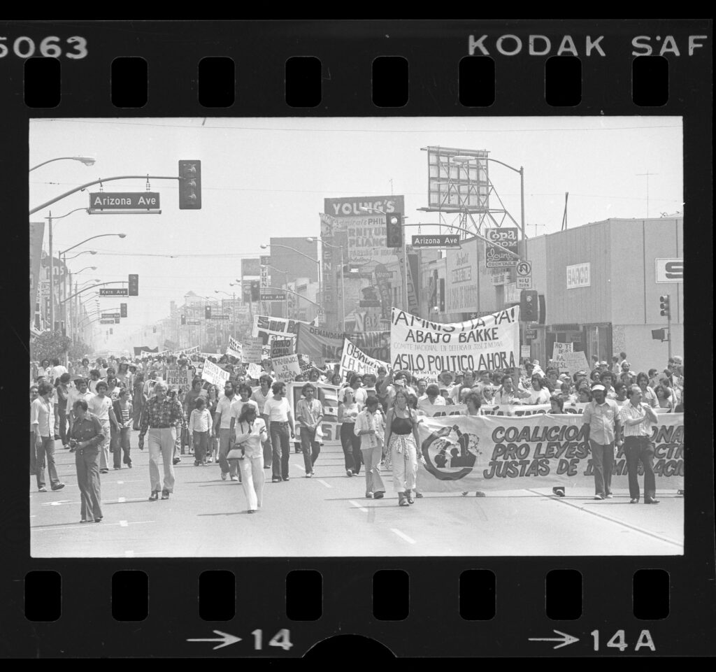 Black and white image appears as a negative with perforated strips on top and bottom. It depicts a large demonstration down a street with a street light and signs reading "Arizona Ave." Marchers carry banners in Spanish. The sign in the front reads "Coalición Pro Leyes y Prácticas Justas de Inmigración." Another, held up high behind it reads " íAmnistia ya! / Abajo Bakke / Comite Nacional In Defensa De José J. Medina" /Asilo Politico Ahora."