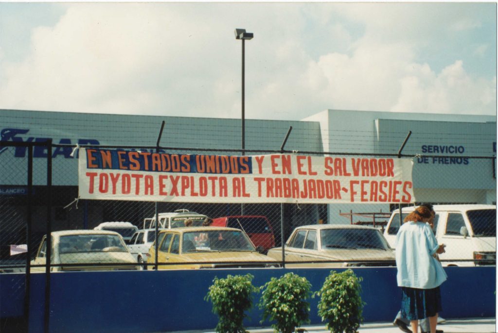 Exterior shot of the Goodyear plant and its parking lot. On the fence dividing the lot from the sidewalk, there's a banner hanging that reads "En Estados Unidos y en El Salvador Toyota Exploit al Trabajador-Feasies" ("Toyota Exploits the Worker in the U.S. and in El Salvador.")